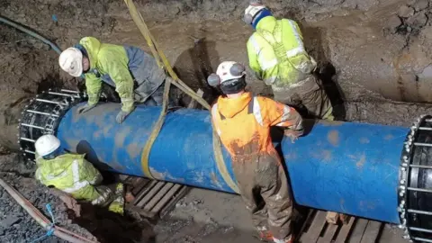 Four engineers wearing muddy fluorescent waterproofs and white helmets as they work to repair the burst water main on Pollokshaws Road in Glasgow. They are in a ditch securing a large blue pipe section, which has the yellow sling used to lower it still attached.