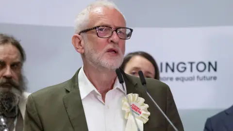 Jeremy Corbyn, wearing a rosette advertising his independent campaign to return to Parliament during his victory speech at the 5 July election