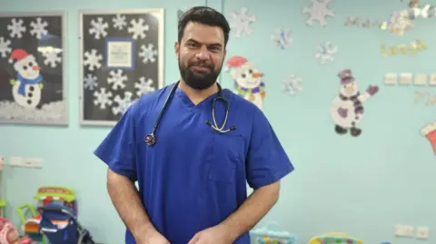 Aisha Iqbal/BBC A man aged in his 30s or early 40s stands smiling. He is South Asian. He wears blue medical scrubs and has a stethoscope hung round his neck. He stands in a children's playroom with walls decorated with Christmas themed pictures, snowmen and snowflakes.