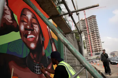 Sodiq Adelakun / REUTERS A man kneeling under scaffolding applies the finishing touches to a mural of a woman wearing a red headscarf and a large grey necklace. Another man looks on while a block of flats can be seen in the background