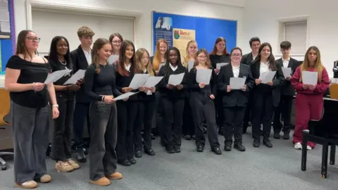 A group of students stands in a classroom or music room, holding sheets of paper and appearing to sing or rehearse. They are wearing black clothes. Behind them, a blue bulletin board displays colourful posters. A piano is visible on the right side