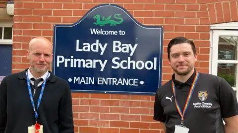 2 men smiling outside Lady Bay Primary School. Man on left in black jumper and tie and man on right in black Notts County Foundation top. 