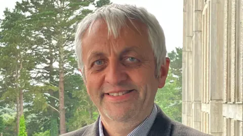 A man with grey hair and a shirt and jacket smiling in front of some trees