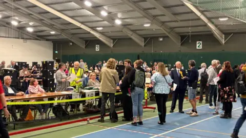 A sports hall with a blue sports court on the floor is filled with people who are sat behind desks with black boxes that are filled with voting slips. A crowd of people are also gathered around in parts of the hall. 