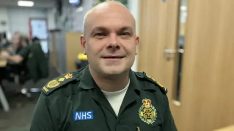 SWAST CEO John Martin stands facing the camera. He is wearing his NHS uniform which is olive green and has yellow badges on it and "NHS" in white letters on a blue patch