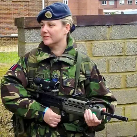 Chelley Tulloch A woman is wearing army fatigues and is holding a very-realistic looking training gun. She is wearing a blue beret and has her blonde hair tied back. 