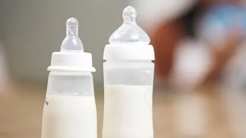 Getty Images Two bottles for feeding babies milk in focus - a mother and baby, out of focus, behind - stock shot
