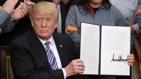 Getty Images President Donald Trump holds up a piece of paper with his signature while surrounded by steel workers in uniform