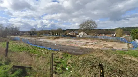 Google The GIS site in Cinderford. There is grass bordering the concrete site which is blocked off by grey fencing and blue netting. There are brown houses behind the site.