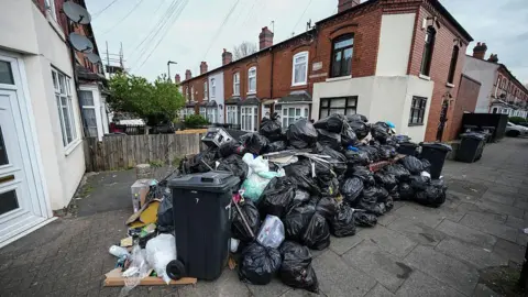 A huge pile of bin bags on a residential street. There are dozens of refuse sacks with terraced homes in the background. 