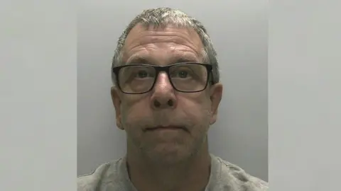 Devon and Cornwall Police mugshot of Peter Wilkinson. He has large black-rimmed glasses on and a grey jumper.