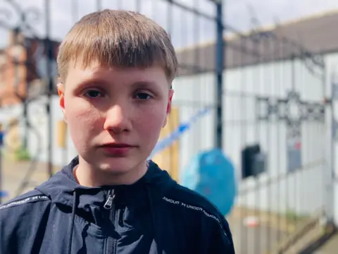 Mason. He is a teenage boy wearing a tracksuit and he has short dark hair. He is standing next to gates.
