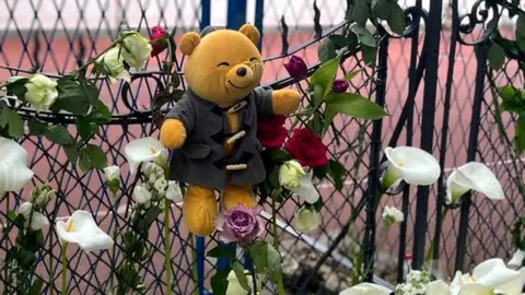 A bear hangs on a fence with white lillies below