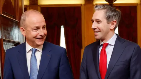 A close up image of Micheál Martin and Simon Harris. On the left, Martin is wearing a navy suit with a blue tie and white collared shirt, and on the left Simon Harris is wearing a dark coloured suit, a red tie and white collared shirt. Both men have their heads toward each other and are smiling.