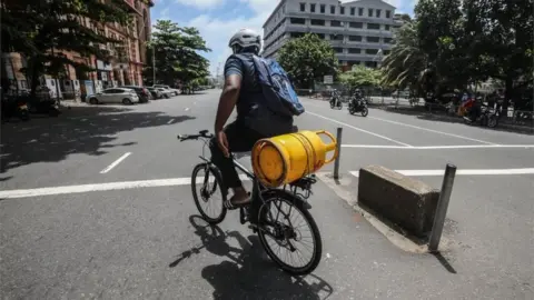 EPA A Sri Lankan man rides a bicycle while he carries an empty cooking gas cylinder on a deserted road amid a fuel shortage in Colombo, Sri Lanka, 29 June 2022