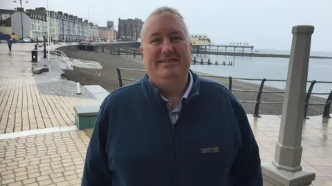 Michael Woods, wearing a navy blue zipped jumper looking at the camera. Aberystwyth prom and the sea are the backdrop. 