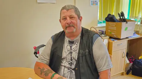 A photo of a white man with grey hair and a goatee beard. He is wearing a grey top with a dark coloured tank top jacket. He is sat at a table with a desk and carboard box in the background
