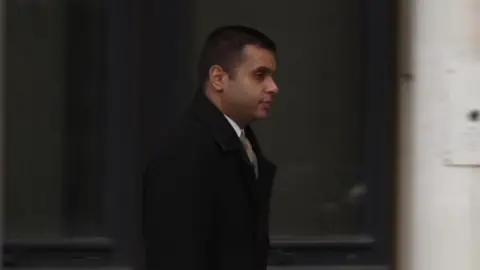 Faisal Shoukat pictured walking away from the court wearing a black overcoat and gold tie. He is carrying a briefcase.