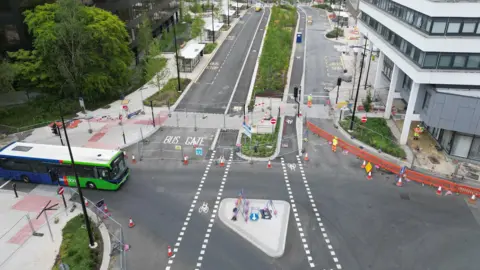 A junction between four roads with fencing, newly planted shrubbery, red cones and new markings for bus stops and cycle paths. A green and blue bus can be seen entering the junction from the left and a few workpeople can be seen on the right wearing hard hats and hi-viz jackets.