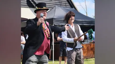 Contributed A man wearing a black top hat, black suit jacket, red T-shirt and grey denim shorts holds a microphone in one hand while his other arm is outstretched. Another man wearing a grey wig, white long-sleeved top, suit waistcoat and cargo shorts stands next to him and is holding a stick with a green rag attached to the end. They are looking to the right of the image. 