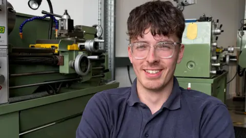 Ben Schofield/BBC Myles Dillon looking straight to camera. He is smiling, showing his teeth, and is wearing clear plastic-framed glasses and a dark blue T-shirt. There is large industrial machinery behind him. 