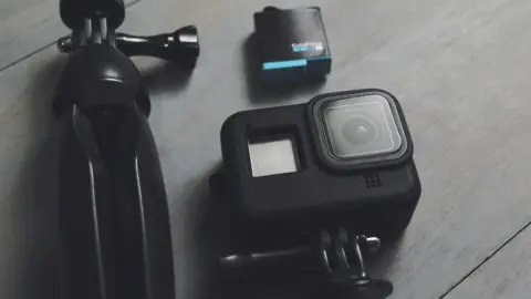 A cube shaped black camera is positioned on a grey wooden surface with a tripod and another piece of equipment that is also black and square.