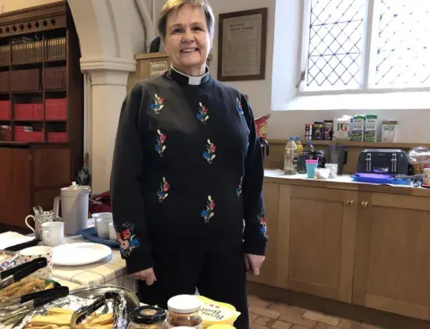 Jon Wright/BBC A female vicar standing inside a church with tea cups and cakes on a table