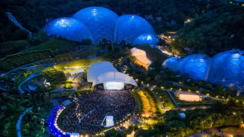 Chris Gorman Eden Project biomes are seen, lit in blue, from above and a crowd is seen in the foreground gathered in front of a stage.
