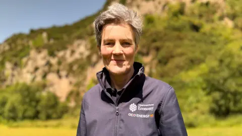 Dr Marie Cowan wears a navy jacket with a Department for the Economy logo on it. She is standing outside in front of a rock face and has short grey hair.