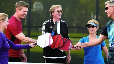 Orange County Register via Getty Images ickleball instructor Mike Fischer, center, congratulates his students after practice on the pickleball courts at Bonita Canyon Sports Park in Newport Beach