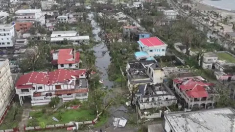 AFP Aerial view of a coastal neighbourhood in Madagascar after a cyclone, showing widespread destruction. Many buildings have damaged roofs, fallen trees block the streets, and floodwater fills the roads. Debris is scattered throughout the area, with the shoreline visible in the background.