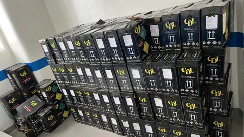 Four rows of black boxes, which are stacked two boxes deep, are lined up against a white will with a blue stripe running along the middle. The boxes have white markings and a yellow logo which reads: "CDL". There are more boxes on in the far left corner of the room.