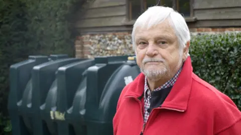 David Coverdale is wearing a red zip-up jumper over a checkered shirt. He has grey hair and a goatee. Behind him is a large dark green oil tank.