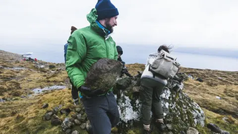 SAVIOUR MIFSUD A Brown Team voluntourist carries a big boulder to be placed on a cairn