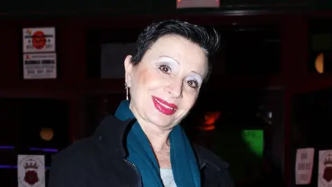 A white woman, with short black hair, tilting her head wearing a blue scarf and black fleece. The background is dark with stickers on the brown wall.