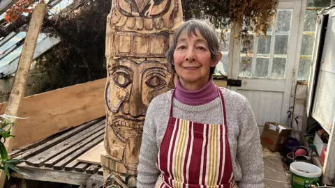 Annabelle Braven stands in front of the wooden pillar she is carving.