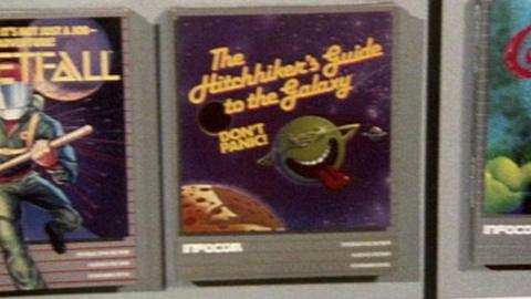 Computer adventure game boxes. The two either side are obscured but the centre one with text on it reading: "The Hitchhiker's Guide to the Galaxy, Don't Panic!".  There is a picture of a planet at the bottom right and a green character with hands over eyes and tongue sticking out.