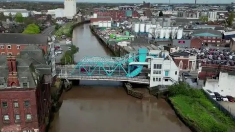 Aerial view of North Bridge. The main blue steel structure spans a muddy river with a large white building on the righthand side. Industrial buildings are visible in the background.