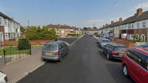 Broad Avenue with the junction to Gloucester Road on the left. It is lined with 1930s terraced houses, with small front gardens and bay windows. There are about seven cars parked along the road and pavement on either side. The road looks recently tarmacked as it is a dark grey. 
