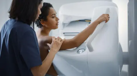 A woman health worker dressed in blue is explaining the mammogram procedure to a patient having a scan. The patient is holding her arm across the machine, and holding on to a rail.