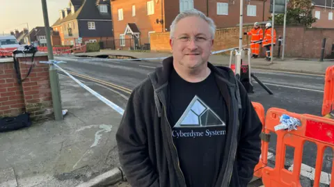 BBC/ADRIAN HARMS Paul Whelan stands in front of the cordoned off sink hole in Godstone. He wears a dark hoody and sweatshirt. Two workmen in hi-vis and safety helmets are standing in the background.