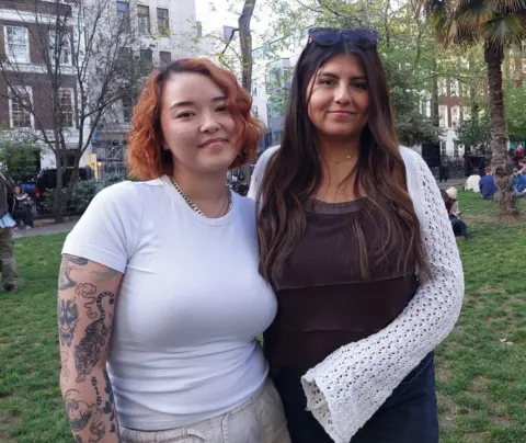 Grace Dean Two women, one with short ginger hair in a white T-shirt and one with long wavy brown hair in a brown and white top, stand up in a park and smile at the camera