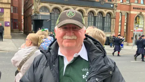 A man wearing a black and green rugby cap wears a black puffer coat and a green rugby jersey. He has white hair and a white moustache and is smiling at the camera.