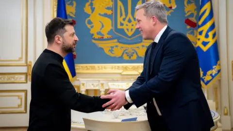 Volodymyr Zelensky, in black mockneck, puts hand forward, which Dan Driscoll, which grey hair and navy suit, grips in both hands. In the background are gold and blue insignia and flags