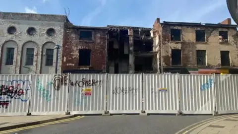 A row of dilapidated buildings on Lower Kirkgate in Leeds, with one partially collapsed. A metal barrier is screening off the row, with the barrier covered in graffiti.  