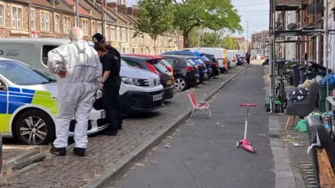 Police officers and forensic officers stand on a residential street in front of a police car.