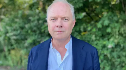 Paul Calvin stands in front of a hedgerow wearing a light blue shirt and dark blue blazer.