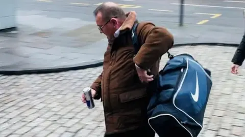 A picture of Rodney Shaw arriving at the High Court in Aberdeen, he is holding a can of juice and is holding a blue sports bag over his shoulder.