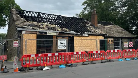 Andrew Jackson/BBC The exterior of the building which housed Kelly's café and Dominoes Hair Designers. The roof has been almost entirely removed by fire. The site has been boarded up with fences around the perimeter.