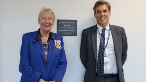 HUC Julie Spence and David Archer stand by a plaque placed on a white wall. Julie is wearing a blue jacket and David a grey jacket, along with a shirt and tie.